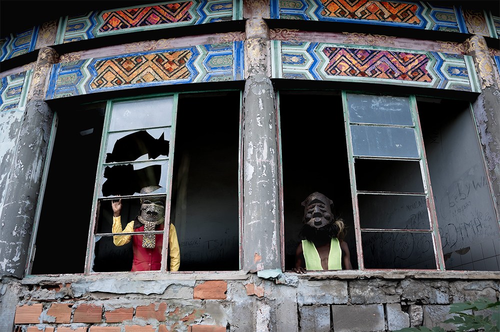 Two people in carved wooden masks look out the broken windows of a ruined building